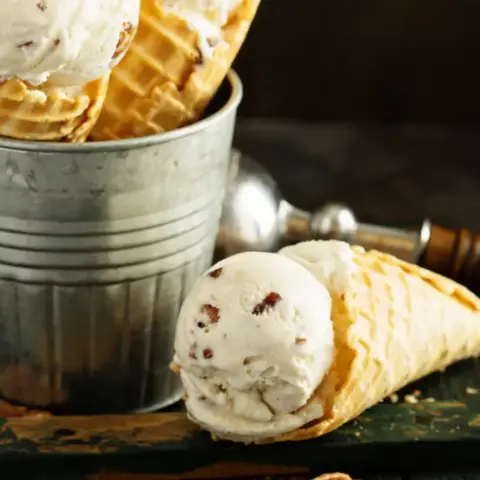 butter pecan ice cream in waffle cone next to bucket with more cones