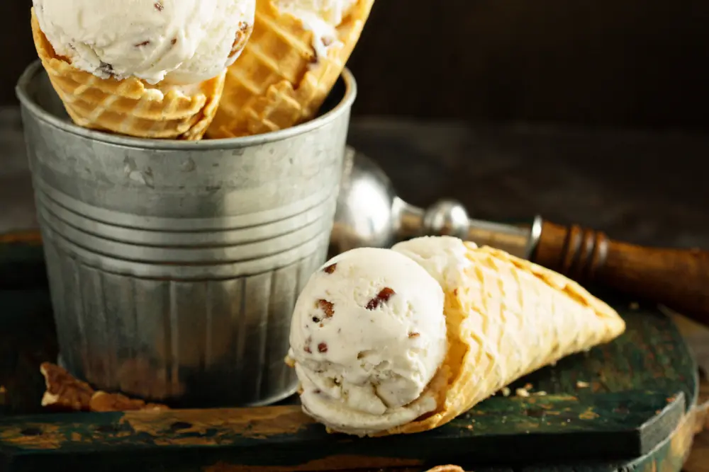 butter pecan ice cream in waffle cone next to bucket with more cones