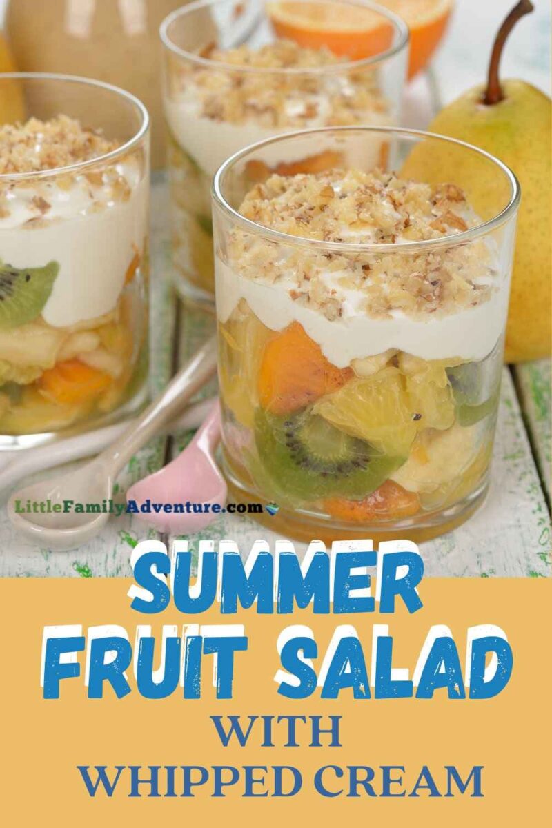 kiwis, pineapple, oranges in glass with whipped cream and nuts on top. Spoons in foreground with words summer fruit salad with whipped cream recipe