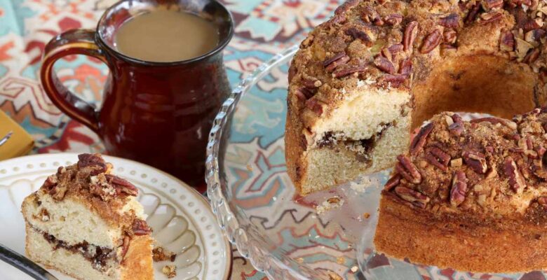slice of german coffee cake on white plate next to brown ceramic mug with a cake plate of a round coffee cake on it