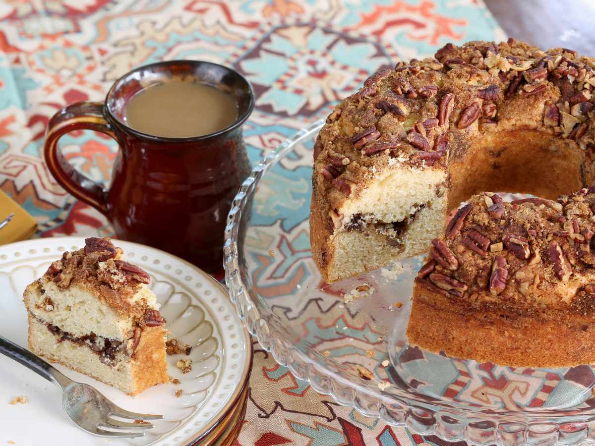 slice of german coffee cake on white plate next to brown ceramic mug with a cake plate of a round coffee cake on it