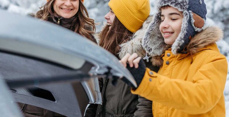 mom with teens bundled in coats and hats next to trunk of car for holiday road trip