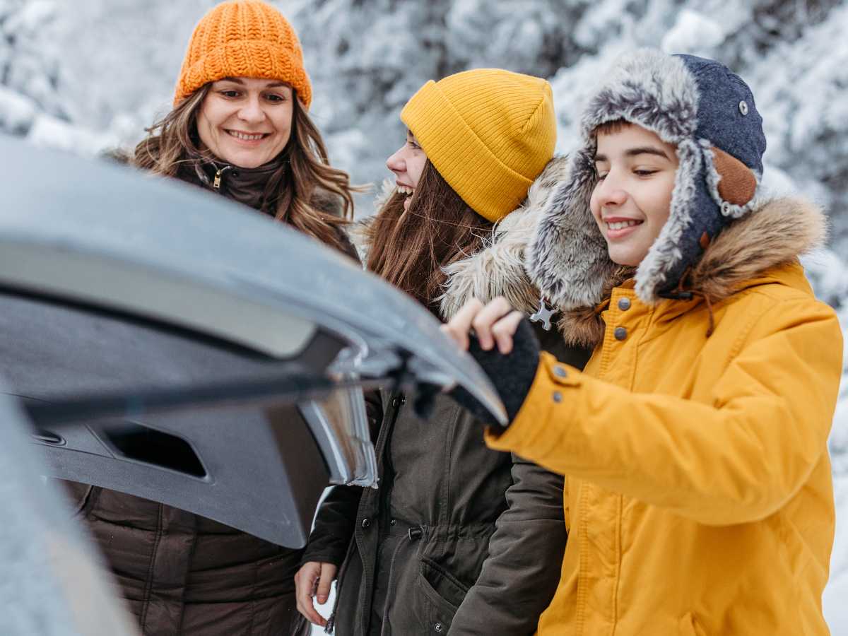 mom with teens bundled in coats and hats next to trunk of car for holiday road trip