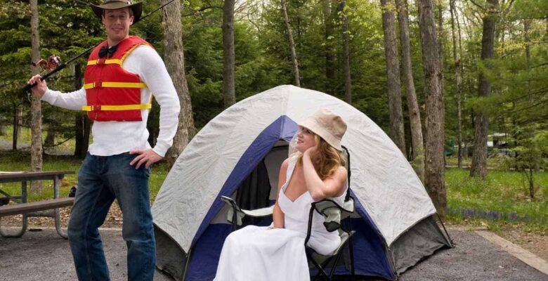 newlyweds in front of tent. husband has life jacket and fishing rod. Bride has wedding dress and hiking boots sitting in a chair