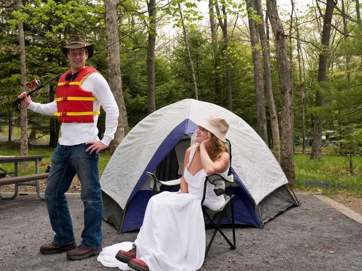 newlyweds in front of tent. husband has life jacket and fishing rod. Bride has wedding dress and hiking boots sitting in a chair