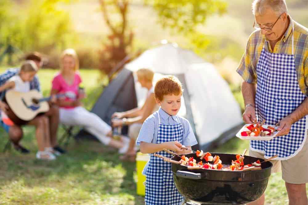 Little boy helping his grampa with easy camping food such as skewers and grilling. Other family and tent in the background