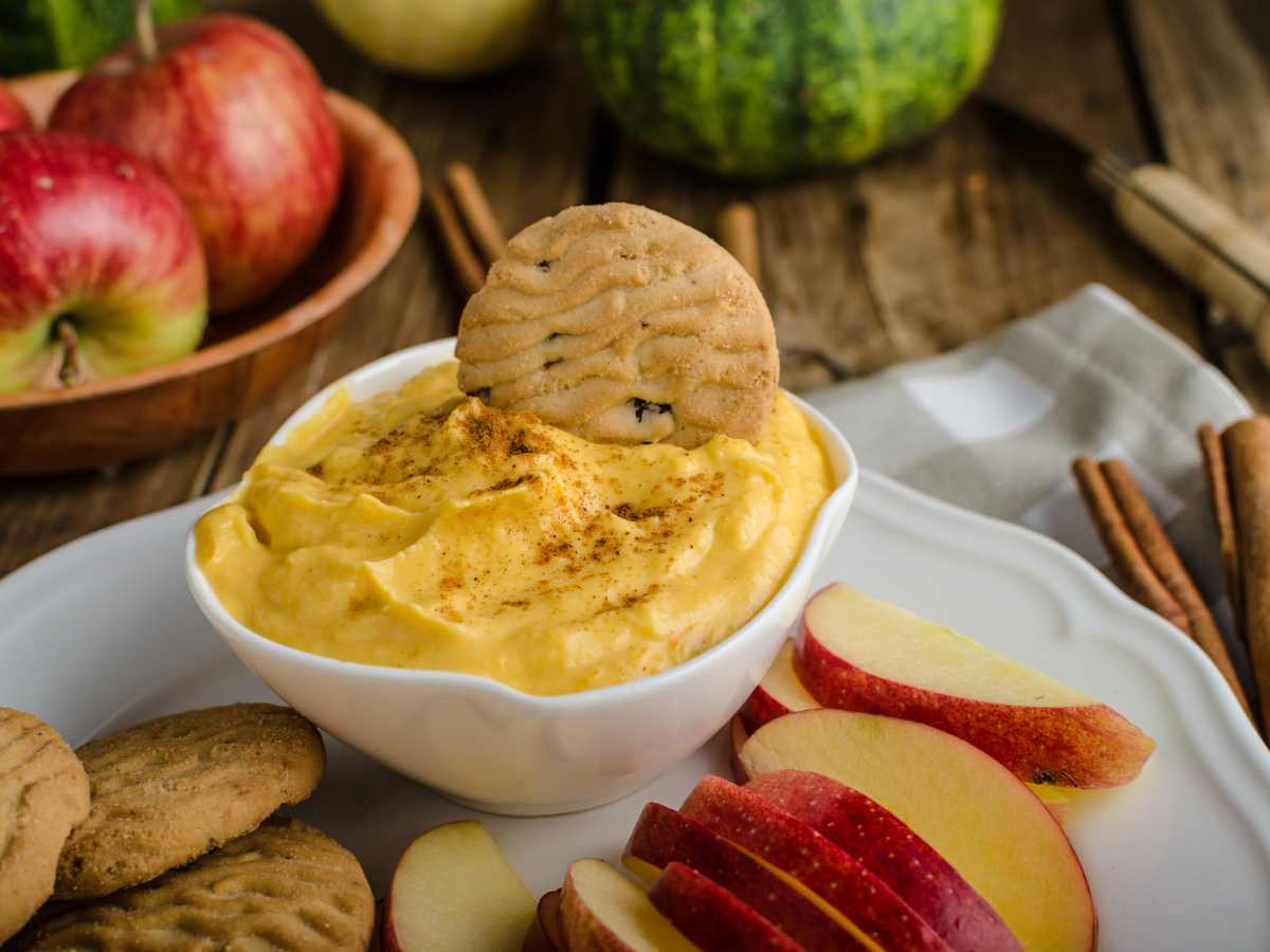 up close photo of 5 ingredient pumpkin pie cheesecake dip in a white ceramic bowl with a thin cookie and apples surrounding it