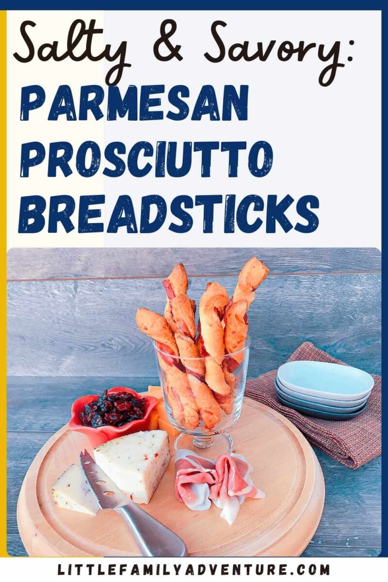 Parmesan prosciutto wrapped breadsticks in glass container on wooden tray with fancy cheeses and crackers