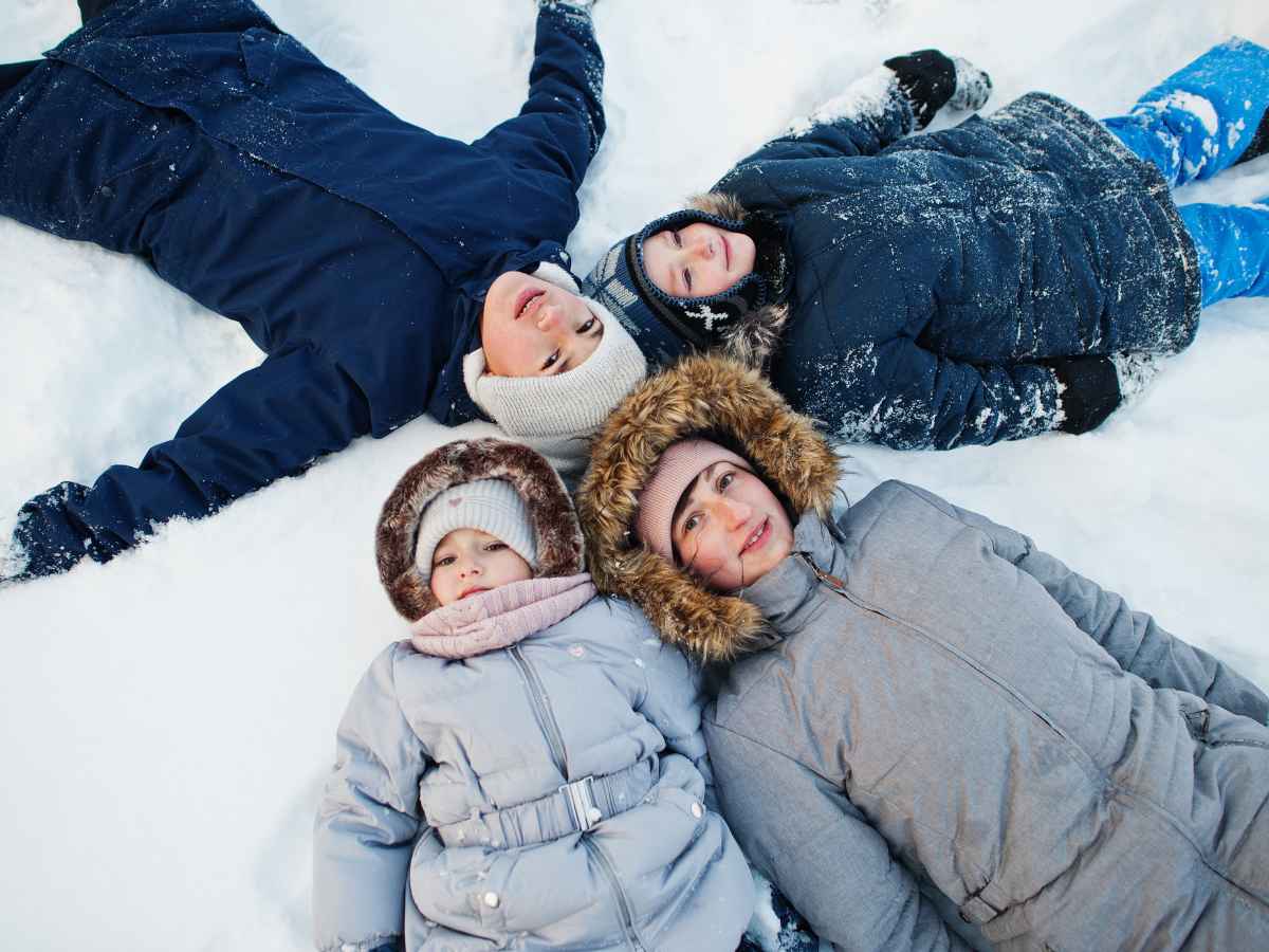 mom and kids in winter coats and hats making snow angels on the ground