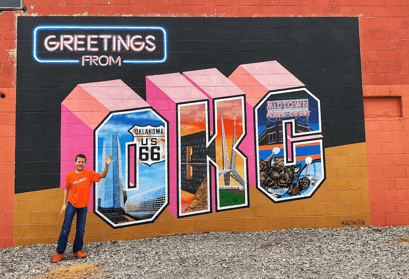 boy waving in front of Greetings from OKC Mural