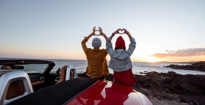 couple sitting on top of car making hearts with their hands at sunset