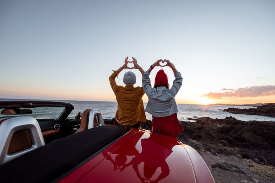 couple sitting on top of car making hearts with their hands at sunset