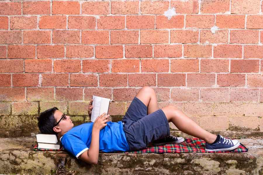teen laying outside by brick wall reading a book