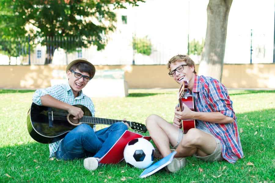 two teen boys sitting in grass with guitar, soccer ball and books
