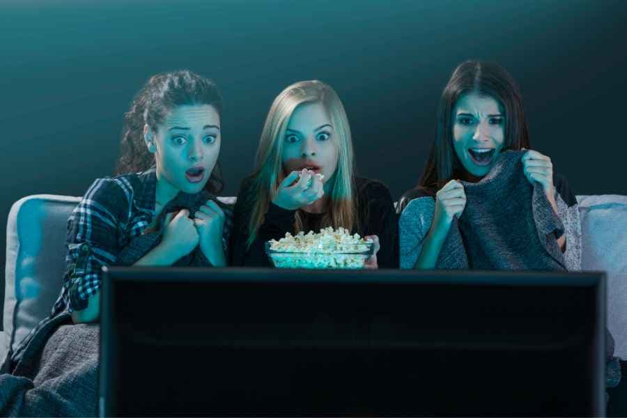 teen girls with frightened expressions eating popcorn and watching a movie in a dark room