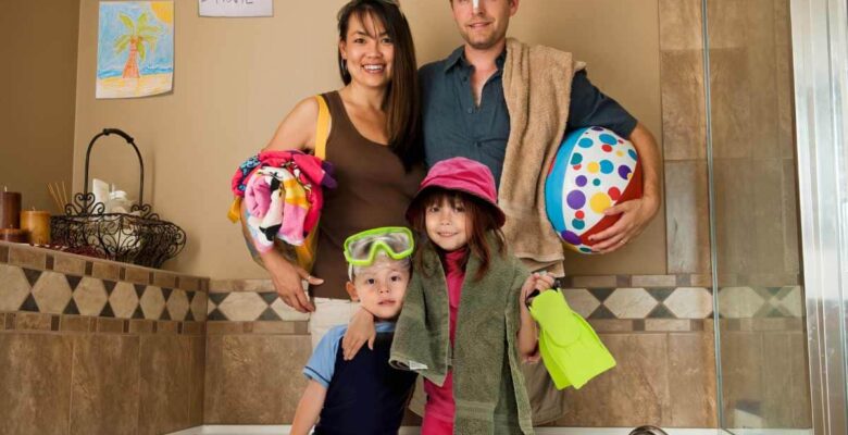 man, woman and two children with beach gear and towels in bathroom with list of staycation ideas hanging on back wall