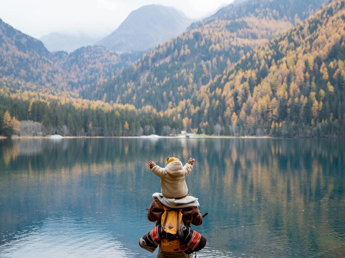 adult with baby on shoulders looking at a mountain lake