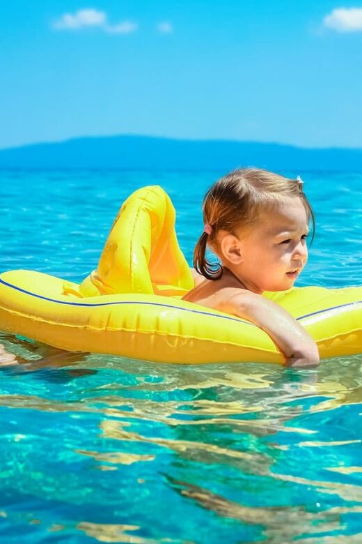 A toddler in floaties standing in clear shallow water at a beach on a bright summer day
