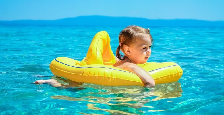 A toddler in floaties standing in clear shallow water at a beach on a bright summer day