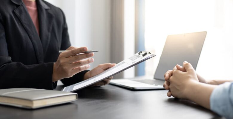 Professional in a dark blazer holds a clipboard and pen while reviewing paperwork with a client seated across a desk with a laptop.